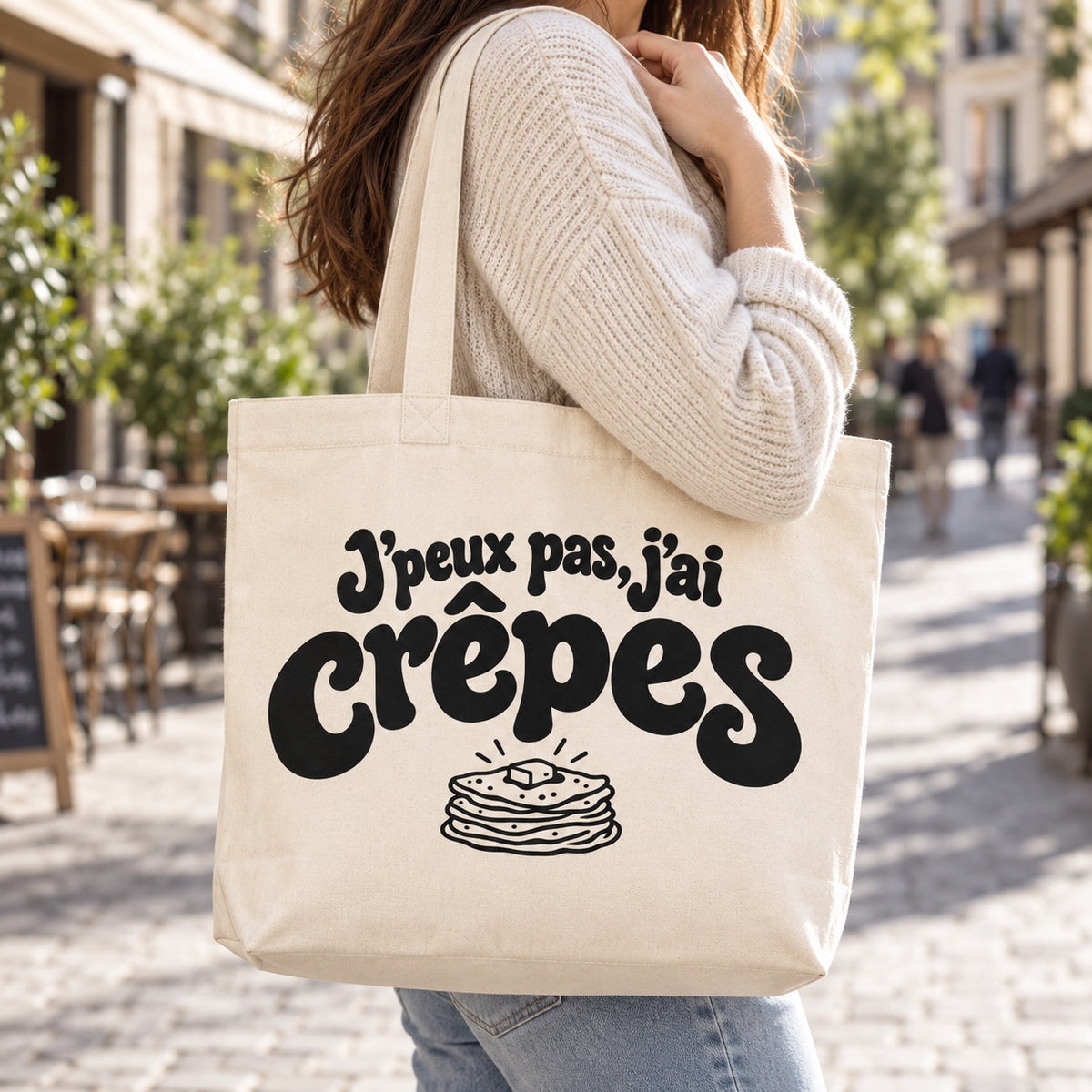 Tote bag « J'peux pas, j'ai crêpes » Premium porté à l'épaule dans une rue piétonne ensoleillée