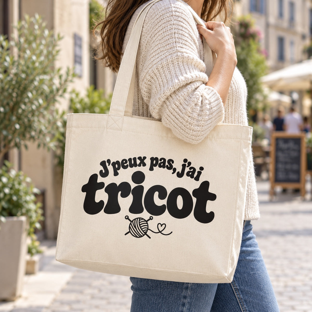 Tote bag « J'peux pas, j'ai tricot » Premium porté à l'épaule dans une rue piétonne ensoleillée