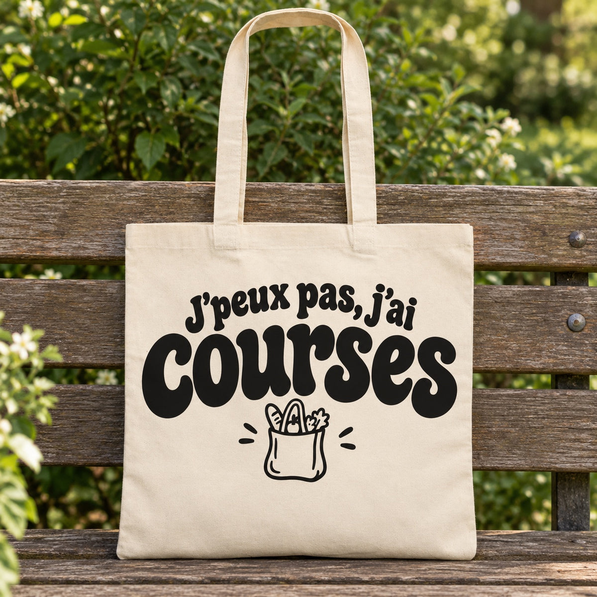Tote bag « J'peux pas, j'ai courses » Essentiel posé sur un banc en bois dans un jardin