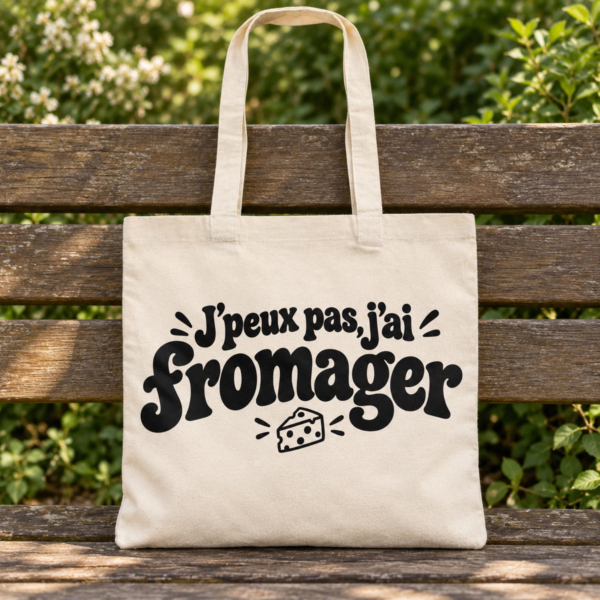 Tote bag « J'peux pas, j'ai fromager » Essentiel posé sur un banc en bois dans un jardin