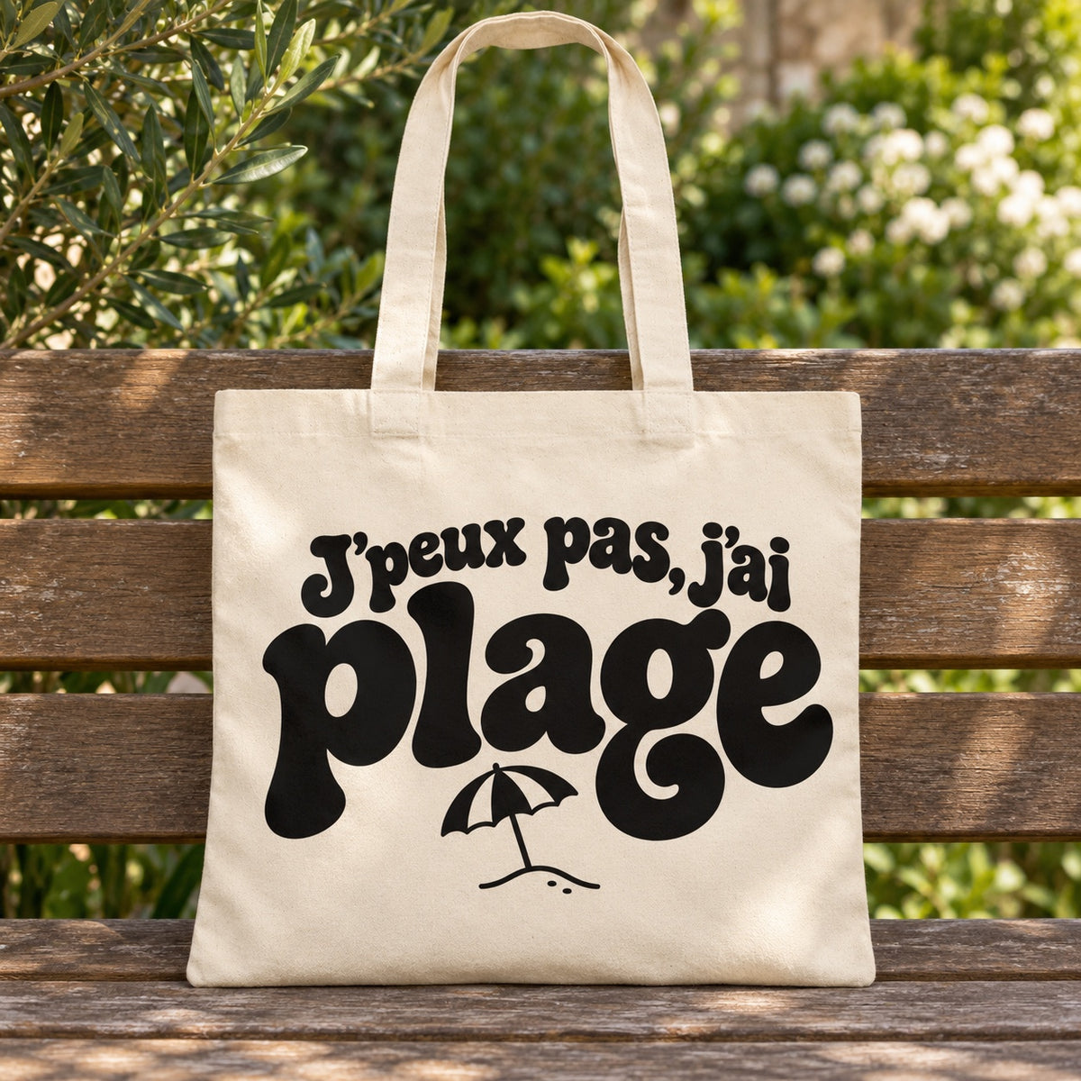Tote bag « J'peux pas, j'ai plage » Essentiel posé sur un banc en bois dans un jardin