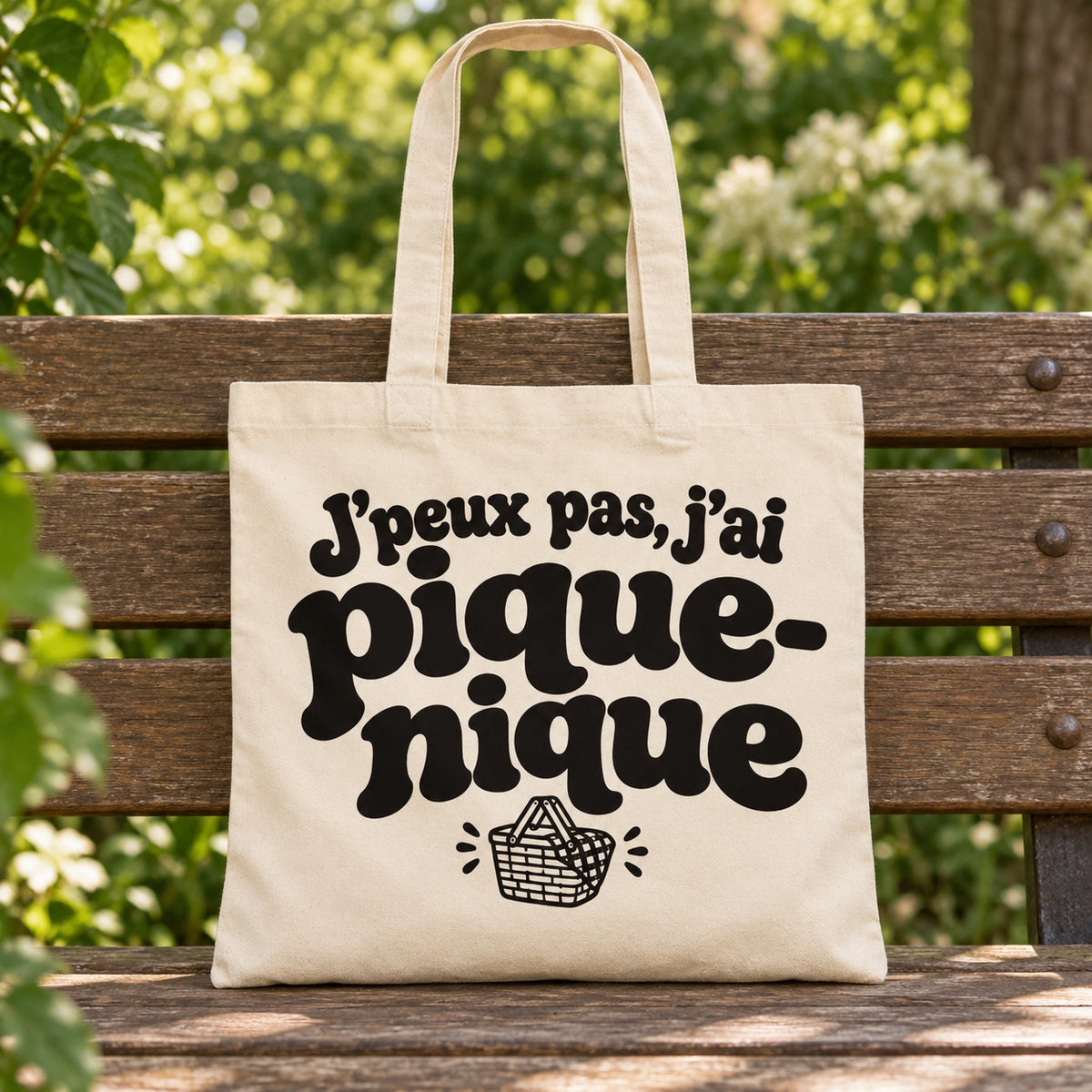 Tote bag « J'peux pas, j'ai pique-nique » Essentiel posé sur un banc en bois dans un jardin