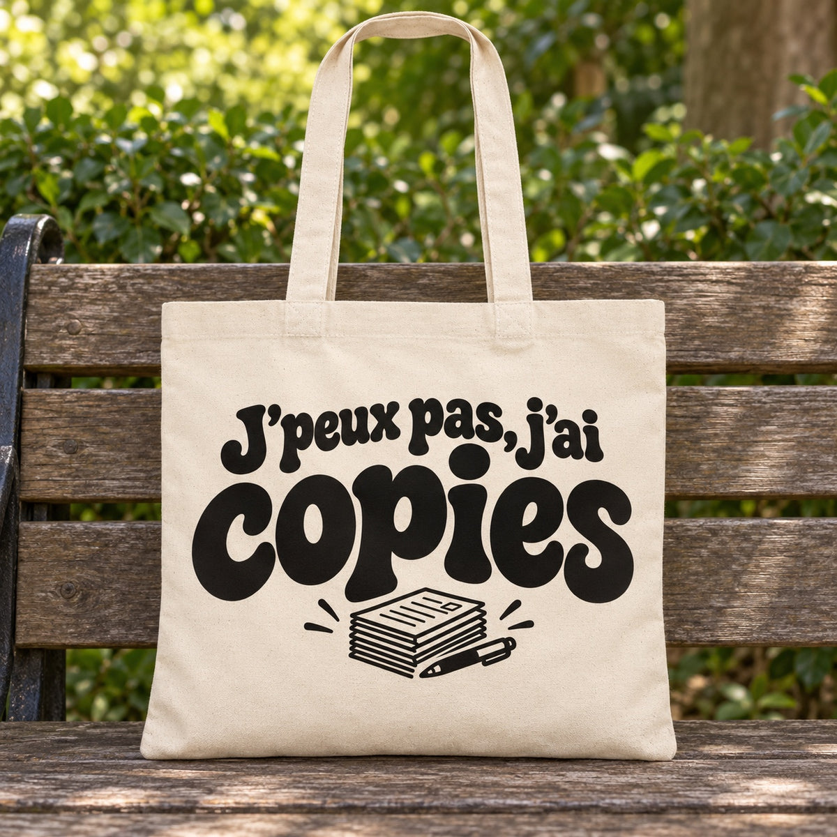 Tote bag « J'peux pas, j'ai copies » Essentiel posé sur un banc en bois dans un jardin