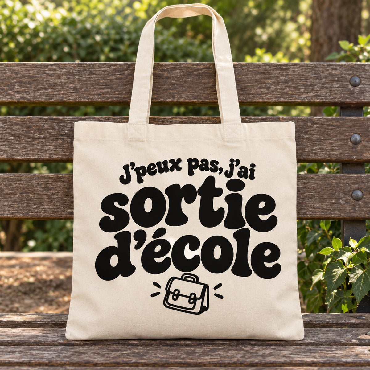 Tote bag « J'peux pas, j'ai sortie d'école » Essentiel posé sur un banc en bois dans un jardin