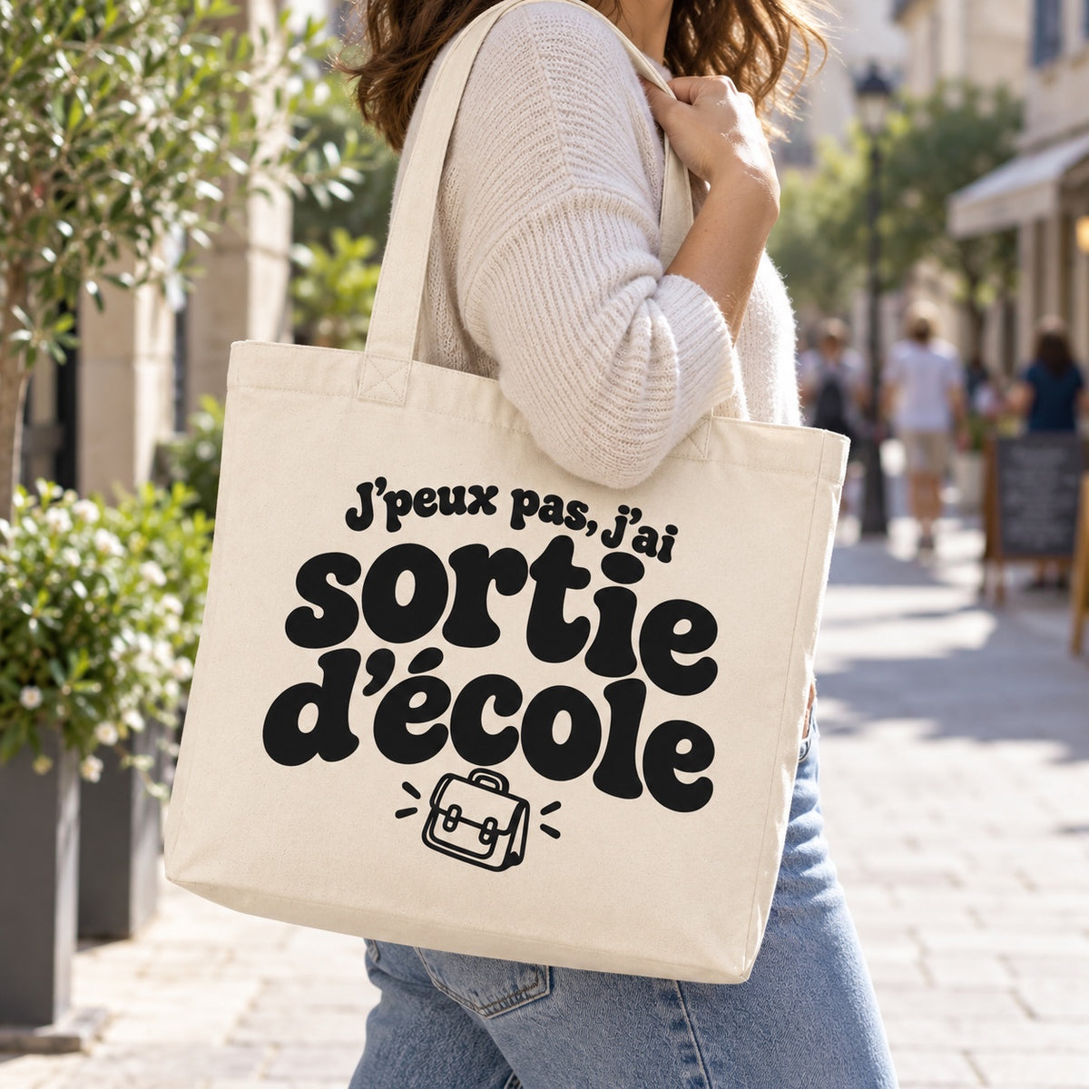 Tote bag « J'peux pas, j'ai sortie d'école » Premium porté à l'épaule dans une rue piétonne ensoleillée