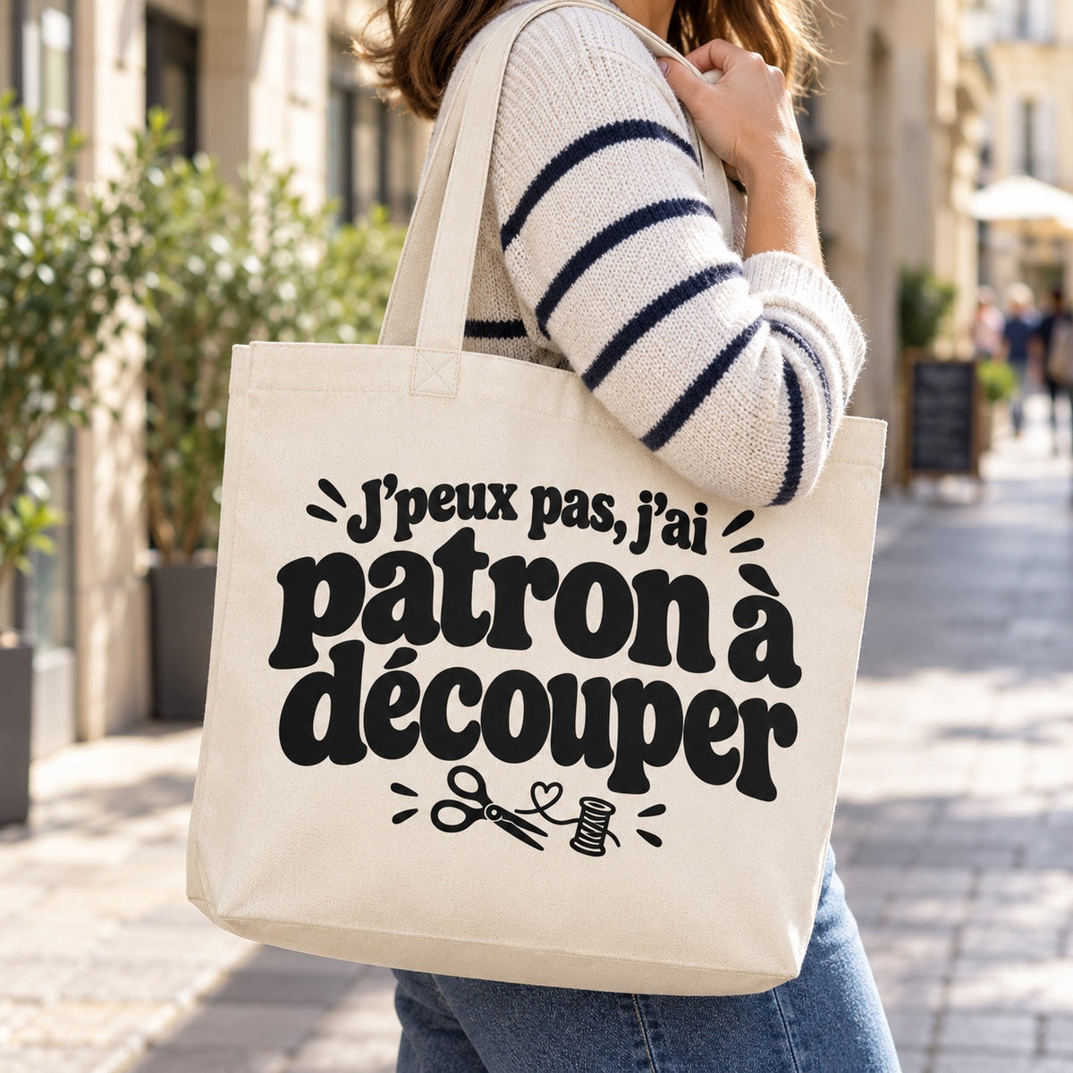 Tote bag « J'peux pas, j'ai patron à découper » Premium porté à l'épaule dans une rue piétonne ensoleillée