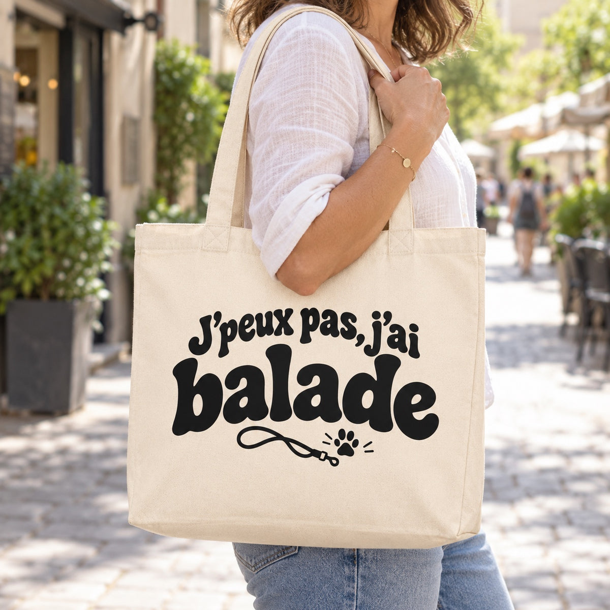 Tote bag « J'peux pas, j'ai balade » Premium porté à l'épaule dans une rue piétonne ensoleillée